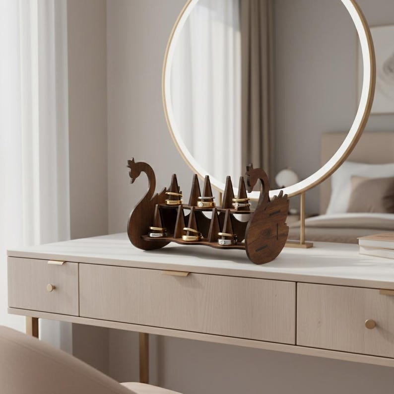 May include: A wooden ring holder shaped like a swan, with multiple ring slots. The holder is dark brown and sits on a light-colored dresser. A round mirror with a gold frame is in the background.