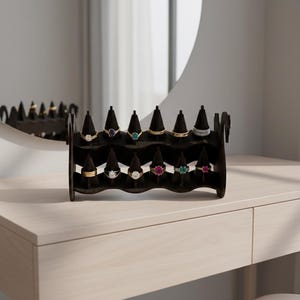 May include: A black ring display stand holding various rings with gemstones. The rings are displayed on black velvet cones. The stand is on a light-colored surface, with a mirror in the background.