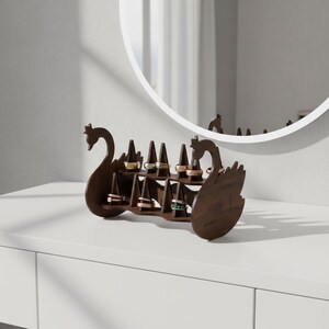 May include: A dark brown wooden ring holder shaped like two swans facing each other. The holder has multiple slots to display rings of various colors and designs. The holder is on a white surface.