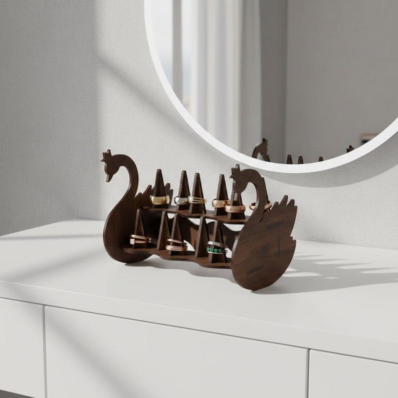 May include: A dark brown wooden ring holder shaped like two swans facing each other. The holder has multiple slots to display rings of various colors and designs. The holder is on a white surface.