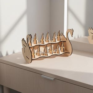 May include: A wooden ring holder in the shape of a swan. The holder has two tiers with multiple slots to display rings. The swan is made of light-colored wood and is sitting on a light-colored surface. The rings are gold and silver.