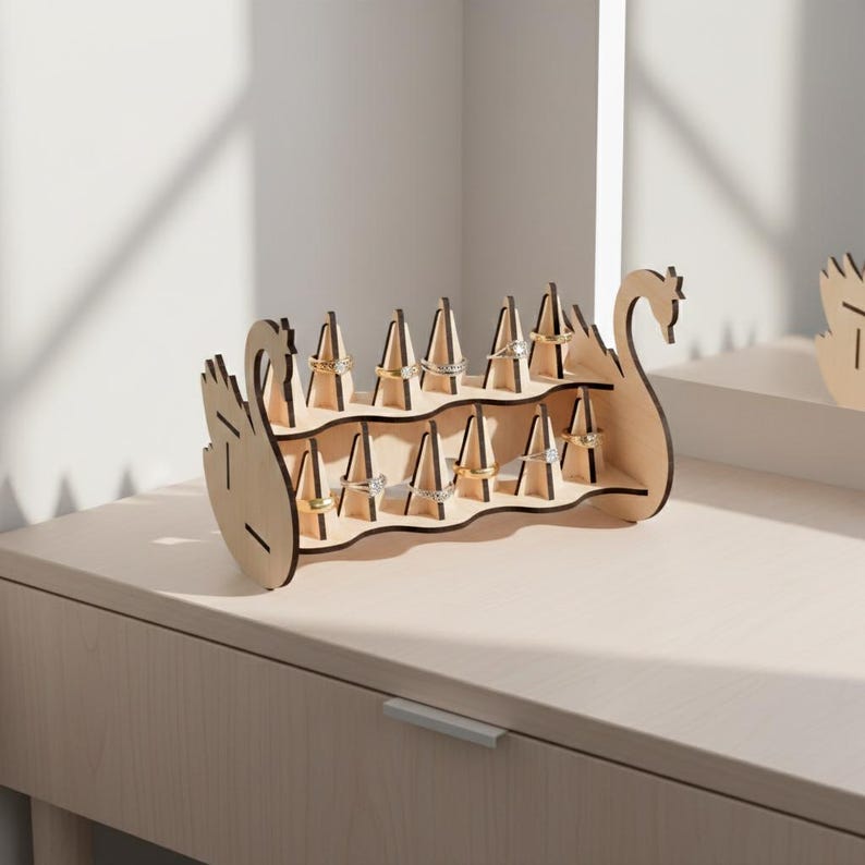 May include: A wooden ring holder in the shape of a swan. The holder has two tiers with multiple slots to display rings. The swan is made of light-colored wood and is sitting on a light-colored surface. The rings are gold and silver.