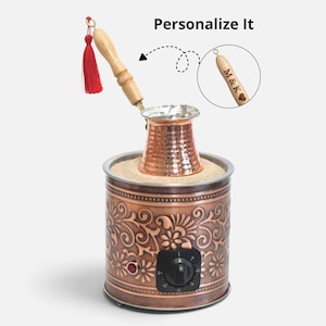 Peut inclure: Une cafetière turque en cuivre avec une finition martelée et un manche en bois clair orné d'un pompon rouge. Elle repose dans un chauffe-café cylindrique en cuivre rempli de sable, décoré de motifs floraux en relief, d'un cadran noir et du texte Personalize It.