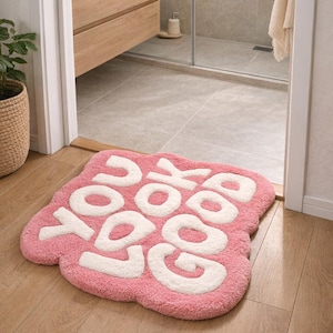 Puede incluir: Una alfombra de baño rosa en forma de nube con las palabras "YOU LOOK GOOD" en blanco, colocada sobre un suelo de madera. La alfombra está en un baño, cerca de un tocador y una ducha.