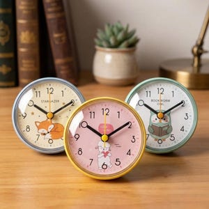 以下が含まれることがあります： 異なる色のフレームと文字盤を持つ、3つの小さな丸いアナログ時計。1つは眠っているキツネ、もう1つは子羊、3つ目は本を読むフクロウです。時計には黒い針が付いています。