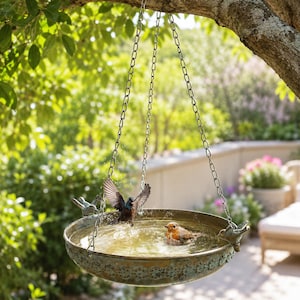 May include: A round, weathered bronze-toned metal bird bath hanging from three silver-colored chains. The basin features a decorative embossed floral pattern along the outer rim and two small bird figurines perched on opposite sides of the edge.