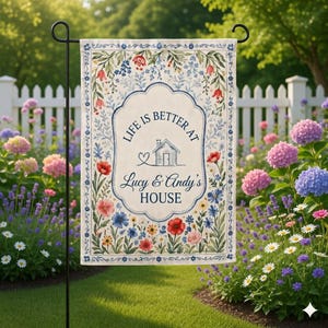 May include: A garden flag with a floral border and the text "LIFE IS BETTER AT Lucy & Andy's HOUSE". The flag features a house graphic and is surrounded by red, blue, and yellow flowers. The flag is displayed in a garden setting.