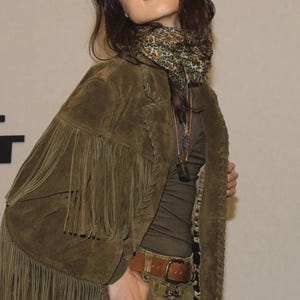 May include: A woman in an olive green suede jacket with fringe, a brown belt, and olive green trousers. She wears a leopard print scarf and a long-sleeved olive green top. A necklace is also visible.