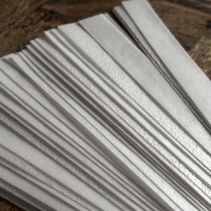 Puede incluir: Una colección de tiras blancas, delgadas y rectangulares dispuestas en forma de abanico. Cada tira presenta una superficie mate y lisa con una textura fina y sutil, creando una apariencia uniforme en toda la pila de piezas individuales.