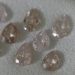 May include: A collection of pear-shaped, light brown diamonds. The gemstones are arranged on a white surface, with facets catching the light. The diamonds vary slightly in color, ranging from pale pink to a deeper brown hue.