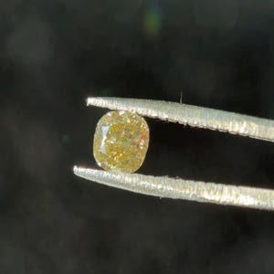 Puede incluir: Un pequeño diamante de talla cojín sujeto con pinzas. La gema tiene un tono amarillo verdoso con inclusiones visibles y una forma ligeramente redondeada. Las pinzas son de color plateado y el fondo es oscuro.