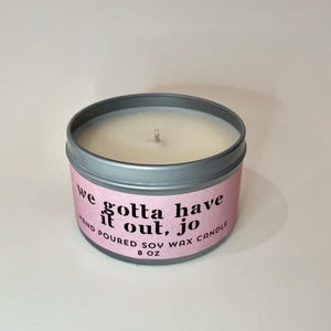 May include: A round, silver-colored candle tin with a pink label. The label reads "we gotta have it out, jo" in black text. The candle is hand-poured soy wax and weighs 8 oz.