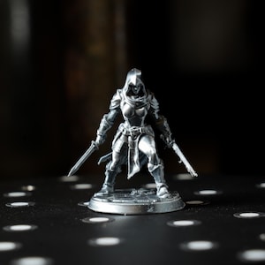 May include: A silver-colored miniature figurine of a warrior woman, clad in armor and a hooded cloak. She is posed with two swords, ready for battle. The figure stands on a circular base, with a dark background.