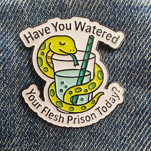 Può includere: Spilla smaltata con bordo bianco, raffigurante un serpente giallo avvolto attorno a un bicchiere d'acqua con una cannuccia verde. Il testo recita "Have You Watered Your Flesh Prison Today?" su sfondo denim.