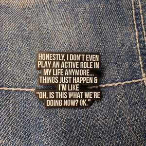 Peut inclure: Épingle en émail noir et blanc avec le texte "HONESTLY, I DON'T EVEN PLAY AN ACTIVE ROLE IN MY LIFE ANYMORE... THINGS JUST HAPPEN & I'M LIKE 'OH, IS THIS WHAT WE'RE DOING NOW? OK.'" sur un fond en jean.