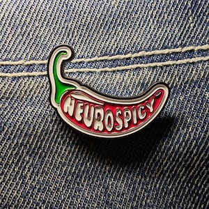 Peut inclure: Une épingle en émail colorée en forme de piment. L'épingle présente un piment rouge avec le mot "NEUROSPICY" en lettres blanches. Le piment a une tige verte et un contour noir, sur un fond en denim.