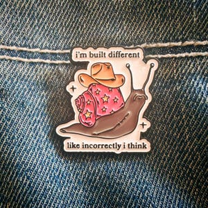May include: A metal enamel pin featuring a brown snail with a pink shell decorated with yellow stars. The snail wears a tan cowboy hat. The text above reads i'm built different and the text below reads like incorrectly i think in black.