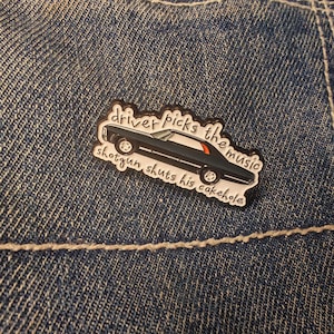 May include: A black car-shaped enamel pin with the text "driver picks the music shotgun shuts his cakehole" in white lettering. The pin has a red stripe on the car's roof and is set against a denim background.