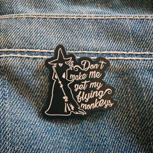 Puede incluir: Un pin de esmalte negro con la silueta blanca de una bruja sosteniendo una escoba. El texto "Don't make me get my flying monkeys" está escrito en una tipografía cursiva blanca a la derecha de la figura, sobre un fondo negro.