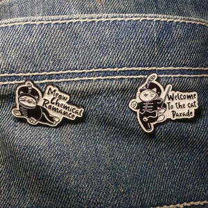 Puede incluir: Dos pines de esmalte con ilustraciones de gatos sobre un fondo de mezclilla. Un pin dice "Meow Chemical Romance" y el otro dice "Welcome To The Cat Parade". Los pines son en blanco y negro con borde blanco.