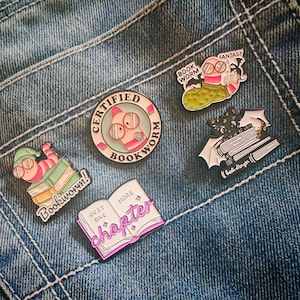 Pode incluir: Quatro pins de esmalte com designs temáticos de livros. Um é um emblema redondo com a inscrição CERTIFIED BOOKWORM. Outros incluem um verme de óculos sobre livros com a etiqueta Bookworm, um verme de suéter com a etiqueta BOOK WORM FANTASY, um morcego sobre livros com a etiqueta book dragon e um livro aberto com o texto JUST ONE MORE chapter.