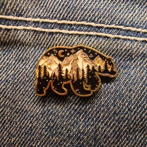 May include: A black and gold bear-shaped enamel pin with a mountain and forest design. The pin features a night sky with a crescent moon and stars. The bear's outline is gold, and the interior is filled with white, black, and gold details.