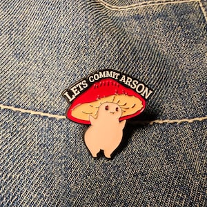 May include: An enamel pin with a cartoon mushroom design and the text "Lets Commit Arson". The mushroom has a red cap and a beige underside, with a smiling face. The pin is set against a blue denim background.