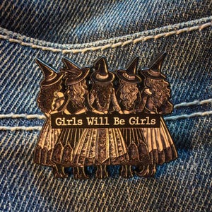 May include: A black and white enamel pin depicting five witches in pointed hats. The pin includes the text "Girls Will Be Girls" on a banner. The pin is set against a blue denim background.