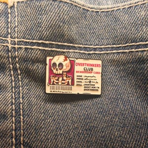 Pode incluir: Um alfinete retangular de esmalte com um design de caveira e esqueleto, com o texto "OVERTHINKERS CLUB MEMBERSHIP CARD". O alfinete está sobre um fundo de ganga azul. O alfinete inclui um código de barras e o texto "ID.NO.17?!".