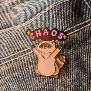 Könnte beinhalten: Ein rosa-brauner Emaille-Pin mit einem Waschbären mit ausgestreckten Armen, unter dem Wort "CHAOS" in rosa Buchstaben. Der Pin hat goldene Akzente und ist vor einem Jeans-Hintergrund platziert.