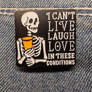 Peut inclure: Épinglette carrée noire avec un squelette blanc tenant une tasse orange. L'épinglette indique "I CAN'T LIVE LAUGH LOVE IN THESE CONDITIONS" en texte blanc. L'épinglette est posée sur un fond en denim.