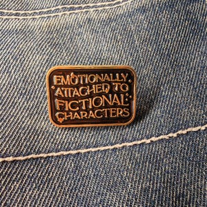 May include: A rectangular enamel pin with a gold border and the text "Emotionally Attached to Fictional Characters" in gold lettering. The pin is set against a blue denim background.