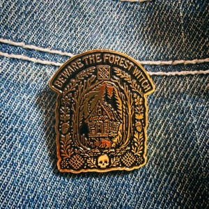 May include: A gold and black enamel pin featuring a cabin in the woods surrounded by floral and acorn motifs. The text BEWARE THE FOREST WITCH is arched across the top, and a small skull is centred at the bottom of the arched frame design.