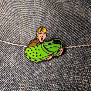 May include: A green Crocs shoe enamel pin with a surprised cartoon character. The pin has black detailing and is set against a denim background. The character is holding the shoe with a look of shock.
