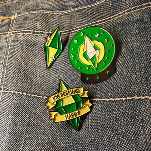 Pode incluir: Três pins de esmalte com um design de gema verde e amarela. Um pin é em forma de gema, outro é uma gema dentro de um círculo verde, e o terceiro tem uma faixa que diz "I'M FEELING HAPPY". Em um fundo de ganga.