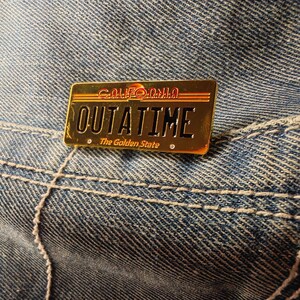 Könnte beinhalten: Eine goldfarbene Anstecknadel in Form eines kalifornischen Nummernschilds. Die Nadel trägt den Schriftzug "CALIFORNIA OUTATIME The Golden State" in einem Retro-Schriftzug. Die Nadel befindet sich auf einem blauen Jeans-Hintergrund.
