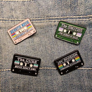 Op de afbeelding: Vier emaille pins in de vorm van cassettes. De pins tonen de namen van bands: Beastie Boys, The Smiths, The Cure en Joy Division. Elke pin heeft een ander kleurenschema en tekst.