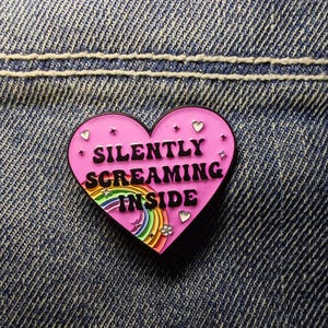 May include: A pink heart-shaped enamel pin with the words "SILENTLY SCREAMING INSIDE" in black lettering. The pin features a rainbow, stars, hearts, and a flower design. The pin is set against a blue denim background.