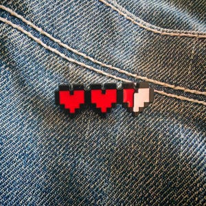May include: A pixelated enamel pin featuring three heart shapes arranged in a horizontal row. The first two hearts are solid red with black outlines, while the third heart is partially red and partially white, creating a segmented pixel effect.