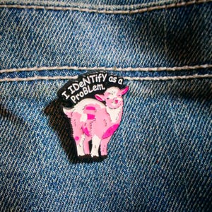 Könnte beinhalten: Eine schwarz-weiße Emaille-Anstecknadel mit einer rosa-weißen Ziege mit verspieltem Ausdruck. Der Text "I IDENTIFY as a ProBLem." steht in weißen Großbuchstaben am oberen Rand der Anstecknadel, die einen schwarzen Umriss aufweist.