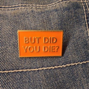 Op de afbeelding: Oranje emaille pin met gouden rand en de tekst "BUT DID YOU DIE?" in gouden letters. De pin is geplaatst tegen een blauwe denim achtergrond met witte stiksels.