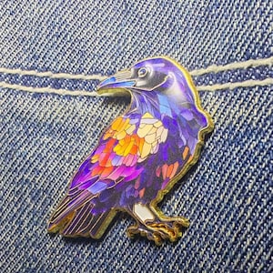 May include: A metal enamel pin shaped like a raven, featuring a vibrant stained-glass style pattern with shades of purple, blue, orange, and yellow. The bird is depicted in profile with gold-toned metal outlines defining its feathers and form.