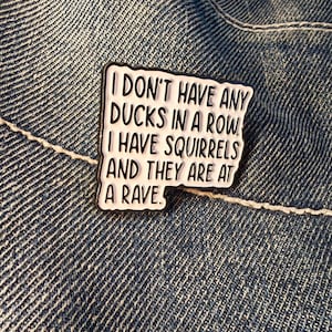 May include: A rectangular enamel pin with a white background and black border. The pin features the text "I DON'T HAVE ANY DUCKS IN A ROW. I HAVE SQUIRRELS AND THEY ARE AT A RAVE." The pin is set against a denim background.