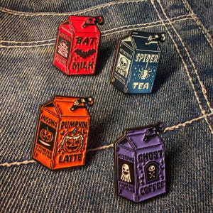 Op de afbeelding: Vier emaille pins in de vorm van melkpakken met Halloween-thema's. De pins zijn rood, blauw, oranje en paars, elk met unieke tekst en afbeeldingen zoals een vleermuis, spin, pompoen en spook. De pins liggen op een denim achtergrond.