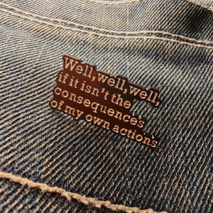 Peut inclure: Épingle en émail de couleur bronze avec l'inscription "Well, well, well, if it isn't the consequences of my own actions" en police serif. L'épingle est posée sur un fond en denim.