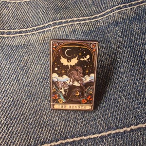 Könnte beinhalten: Rechteckiger Emaille-Pin mit einer Frau, die unter einem Mond und Sternen liest, mit Drachen im Hintergrund. Der Pin hat einen floralen Rand und den Text "THE READER" unten. Auf blauem Jeansstoff.
