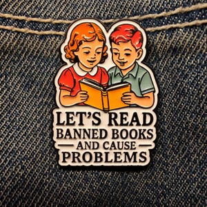May include: A colorful enamel pin featuring two children reading a book. The pin has the text "LET'S READ BANNED BOOKS - AND CAUSE PROBLEMS" in black letters. The pin is set against a denim background.
