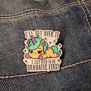 May include: An enamel pin with a cartoon unicorn design in yellow, green, pink, and purple. The pin has the text "I'LL GET OVER IT" and "I JUST NEED TO BE DRAMATIC FIRST". The pin is set against a denim background.