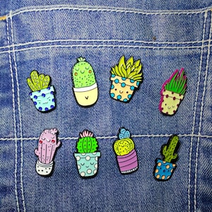 May include: Seven colourful cactus enamel pins. Each pin depicts a unique cactus design in a pot, with various colours including green, pink, yellow, and blue. The pins are set against a blue denim background.