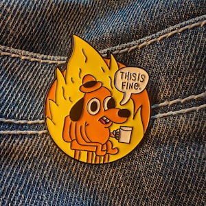 Pode incluir: Um pin redondo de esmalte com um cão de desenho animado sentado numa cadeira, rodeado por chamas. O cão é laranja com chapéu e orelhas pretas, segurando uma caneca. Um balão de fala diz: "THIS IS FINE."